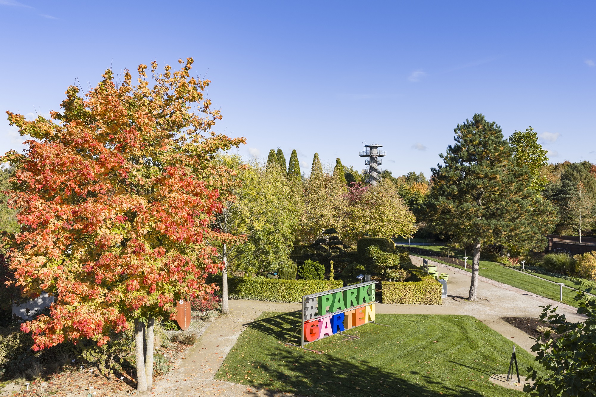 Ein Park mit grünen Rasenflächen, bunten Herbstbäumen und einem großen #PARKGARTEN-Schild in leuchtenden Buchstaben. Wege schlängeln sich durch das Gelände, und im Hintergrund ist ein hoher Aussichtsturm unter einem klaren blauen Himmel zu sehen.