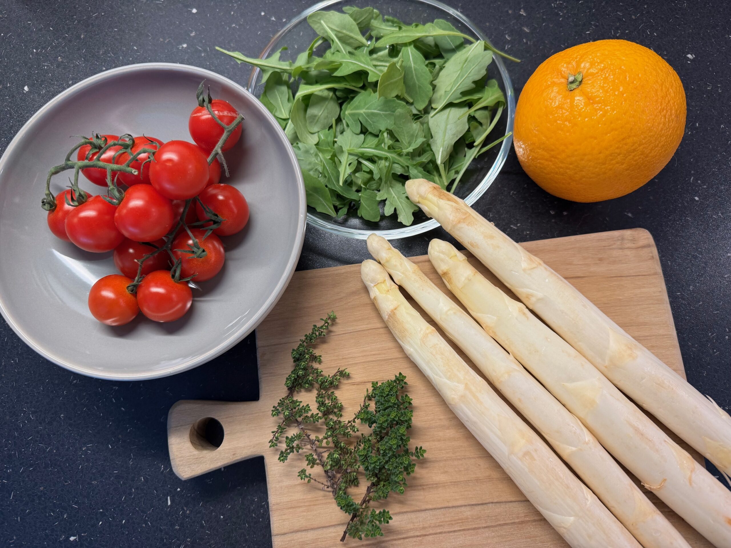 Eine Schale mit Kirschtomaten, eine Schale mit Rucola, eine Orange, weiße Spargelstangen auf einem Holzbrett und frische Thymianzweige stehen auf einer schwarzen Arbeitsplatte - perfekt, um sie mit Ihren Lieblingsrezepten von Brot Backen zu kombinieren.