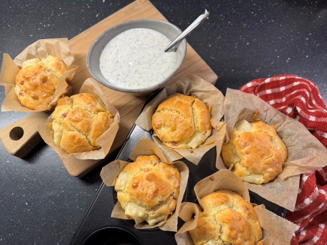 Sechs goldbraune Muffins in Pergamenthüllen auf einem Holzbrett, daneben eine Schale mit cremigem Dip und ein Löffel. Perfekt, um neue Brot Backen Rezepte auszuprobieren. Dieses Arrangement steht neben einem rot-weiß gestreiften Tuch auf einer schwarzen Arbeitsplatte.