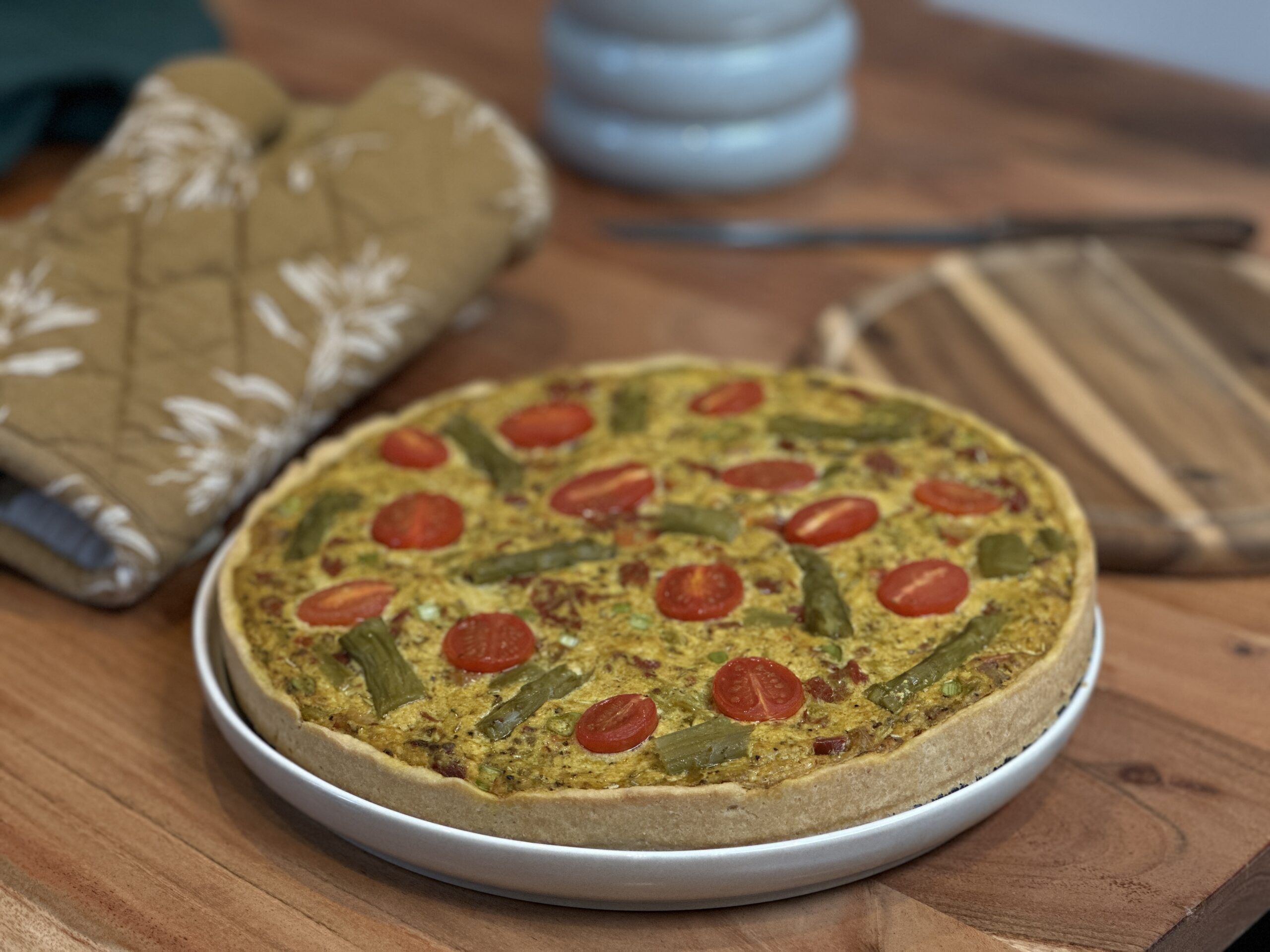 Eine frisch gebackene Quiche mit Kirschtomatenhälften und grünem Spargel liegt auf einem weißen Teller auf einem Holztisch, umgeben von Küchenutensilien - perfekt für alle, die Brot Backen Rezepte und herzhafte Backwaren lieben.