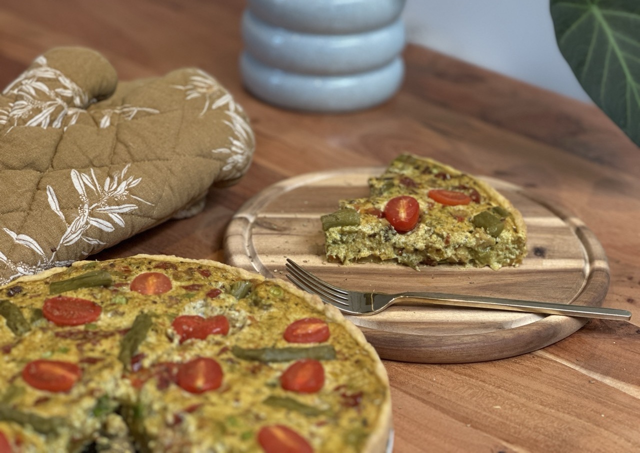Eine Gemüse-Quiche mit Kirschtomaten und grünen Bohnen liegt auf einem Holzbrett, eine Scheibe daneben, eine Gabel daneben - perfekt für die Brot Backen Rezepte. Ein Topfhandschuh und eine Pflanze schmücken den Holztisch.