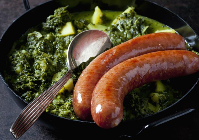 Eine Nahaufnahme von zwei gekochten Würsten, die auf gedünstetem Gemüse und gewürfelten Kartoffeln ruhen, mit einem Servierlöffel aus Metall in der Pfanne - ein herzhaftes Gericht, das perfekt zu Ihren Lieblings-Brotbacken-Rezepten passt.