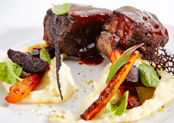 Zwei Stücke glasiertes Rindfleisch auf cremigem Kartoffelpüree, garniert mit gerösteten Karotten, Roter Bete, frischem Grün und einem dünnen, dunklen Knusperstück - perfekt angerichtet für alle, die kunstvolle Gerichte lieben oder neben Gourmetgerichten gerne Brot Backen Rezepte erkunden.