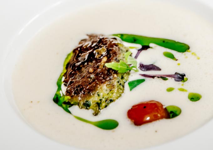 Ein Gourmet-Gericht mit einem gebratenen Patty in Kräuterkruste und einer cremigen weißen Soße, garniert mit grünem Öl, Mikrogrün und einer gerösteten Kirschtomate - perfekt mit frischem Brot aus Ihrem Lieblings-Brotbacken-Rezept.