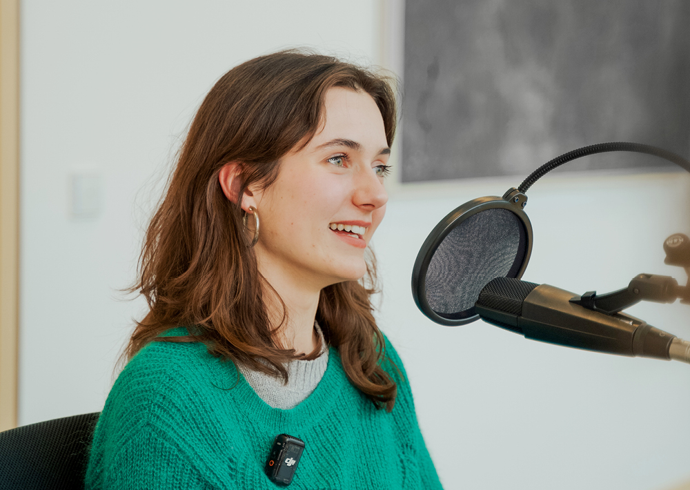 Eine junge Frau namens Marie Becker mit braunen Haaren und grünem Pullover lächelt, während sie in einem Tonstudio in ein Mikrofon spricht. Sie bereitet sich auf ihre Rolle als Marie Becker in der ZDF-Theaterproduktion Ku'damm 77 vor.