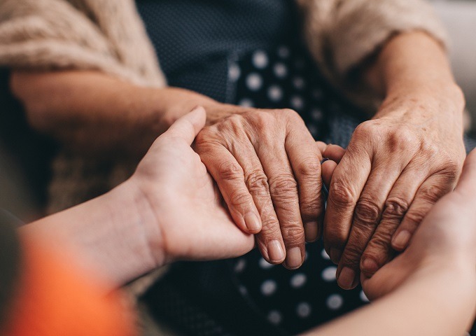 Eine Nahaufnahme einer jüngeren Person, die sanft die Hände eines älteren Erwachsenen hält und so Fürsorge und Unterstützung vermittelt. Die ältere Person trägt ein gepunktetes Kleid und einen beigen Pullover.