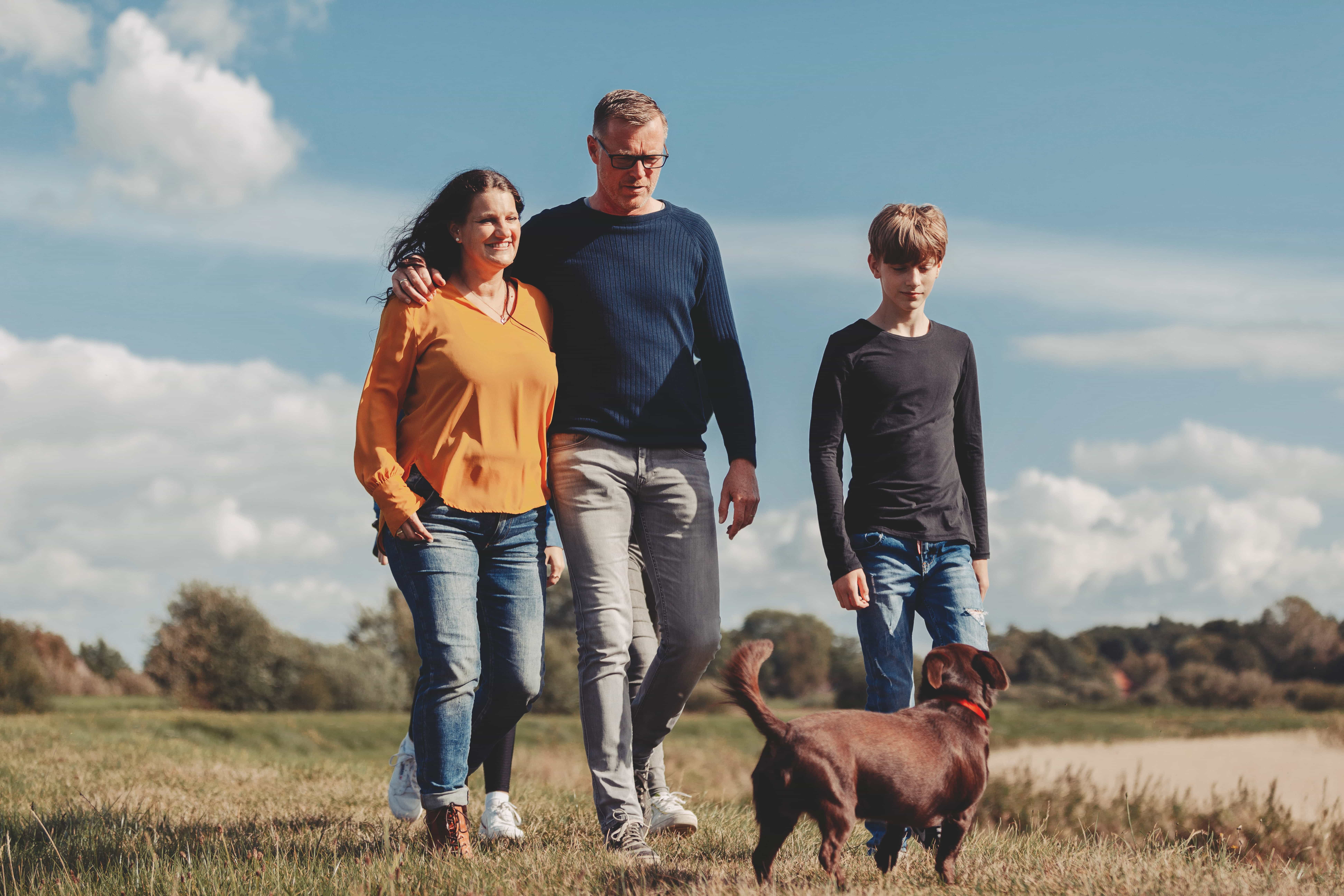 Eine Frau, ein Mann und ein Junge gehen zusammen auf einem grasbewachsenen Weg mit einem braunen Hund. Der Himmel ist blau mit einigen Wolken und Bäumen im Hintergrund. Nähe macht mehr möglich, während sie entspannte Momente zusammen genießen, lässig gekleidet.