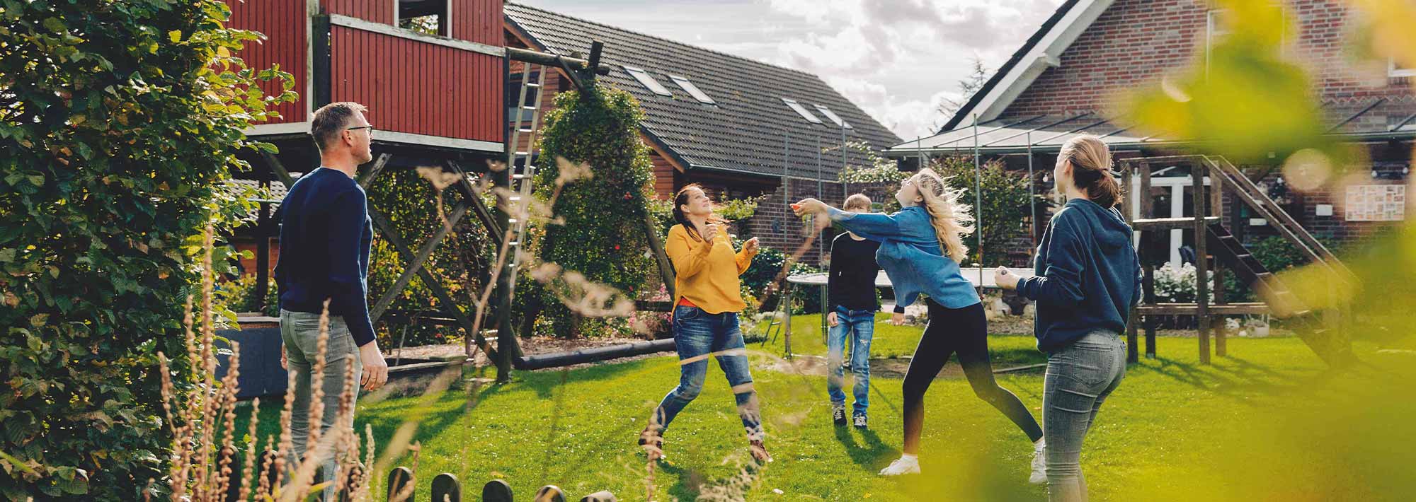 Eine Gruppe von fünf Personen spielt Badminton in einem grasbewachsenen Hinterhof, umgeben von Bäumen und Häusern, mit einem roten, scheunenartigen Gebäude im Hintergrund an einem sonnigen Tag - ein echter Zurück-zum-Anfang-Moment.