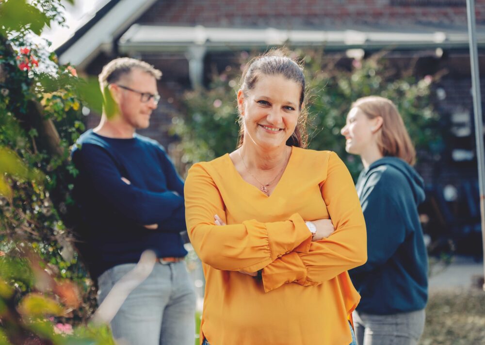 Eine Frau in einer gelben Bluse lächelt mit verschränkten Armen im Vordergrund und strahlt die Wärme von "Nähe macht mehr möglich" aus, während ein Mann und eine andere Frau im unscharfen Hintergrund draußen in der Nähe eines Hauses miteinander plaudern.