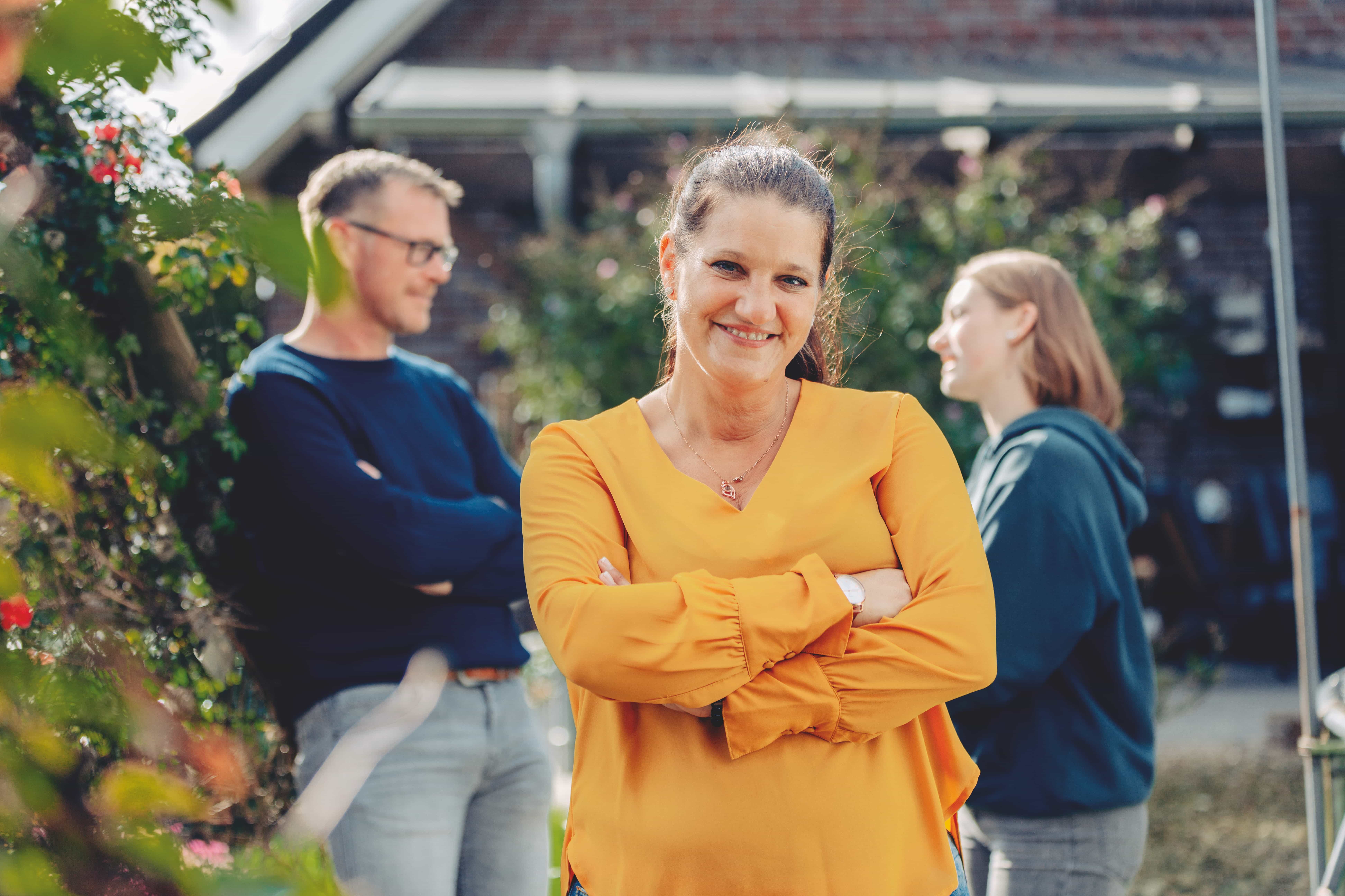 Eine Frau in einer gelben Bluse lächelt mit verschränkten Armen im Vordergrund und strahlt die Wärme von "Nähe macht mehr möglich" aus, während ein Mann und eine andere Frau im unscharfen Hintergrund draußen in der Nähe eines Hauses miteinander plaudern.