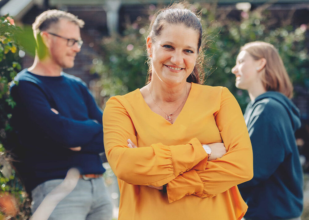 Eine Frau in einer gelben Bluse steht lächelnd mit verschränkten Armen im Vordergrund, während ein Mann und eine andere Frau draußen im Hintergrund stehen und sich unterhalten - ein Moment des Zurück zum Anfang.