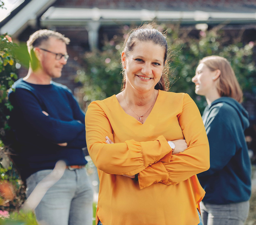 Eine Frau in einer gelben Bluse steht lächelnd mit verschränkten Armen im Vordergrund, während ein Mann und eine andere Frau draußen im Hintergrund stehen und sich unterhalten - ein Moment des Zurück zum Anfang.