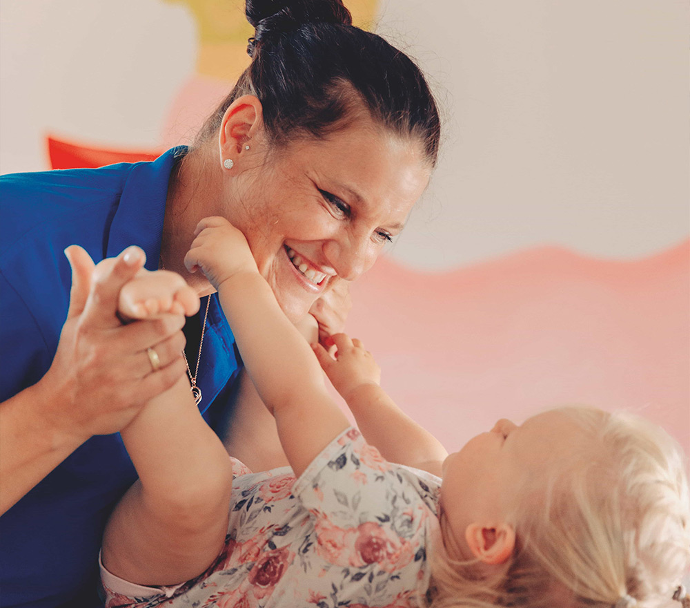 Eine Frau in einem blauen Hemd lächelt warm, während sie mit einem kleinen Kind in einem geblümten Kleid spielt und Händchen hält. Sie schauen sich liebevoll an, als ob sie einen Moment der Freude - "Zurück zum Anfang" - in einer hellen, fröhlichen Umgebung genießen würden.