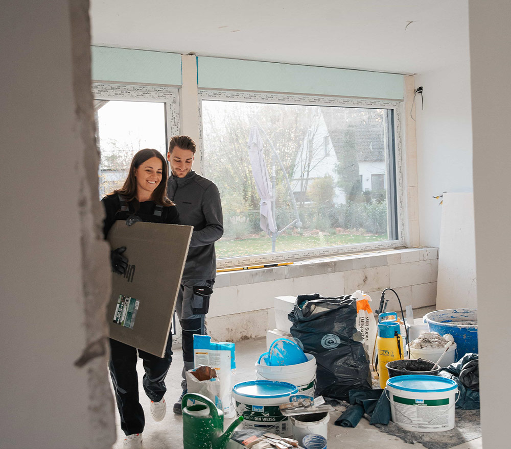 Eine Frau und ein Mann tragen Renovierungsmaterial in einen hellen, teilweise renovierten Raum mit großen Fenstern, Eimern, Werkzeugen und Vorräten, die auf dem Boden verstreut sind - bereit für einen Neuanfang, zurück zum Anfang.
