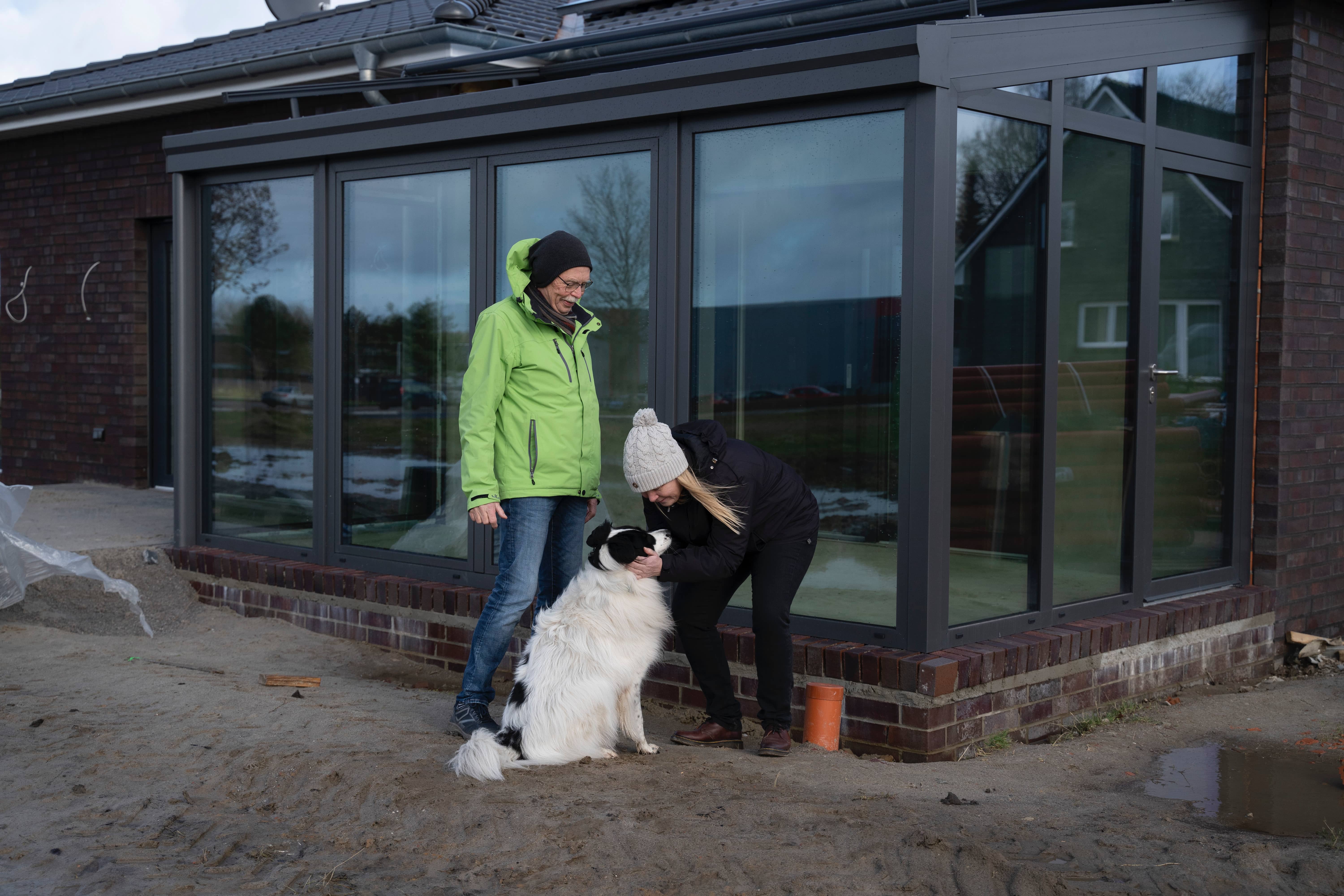 Zwei Menschen in Winterkleidung lächeln und streicheln einen großen schwarz-weißen Hund vor einem modernen Haus mit Glaswänden. Der sandige Boden deutet auf eine neue Konstruktion hin - Nähe macht mehr möglich, während sie diesen besonderen Moment gemeinsam genießen.