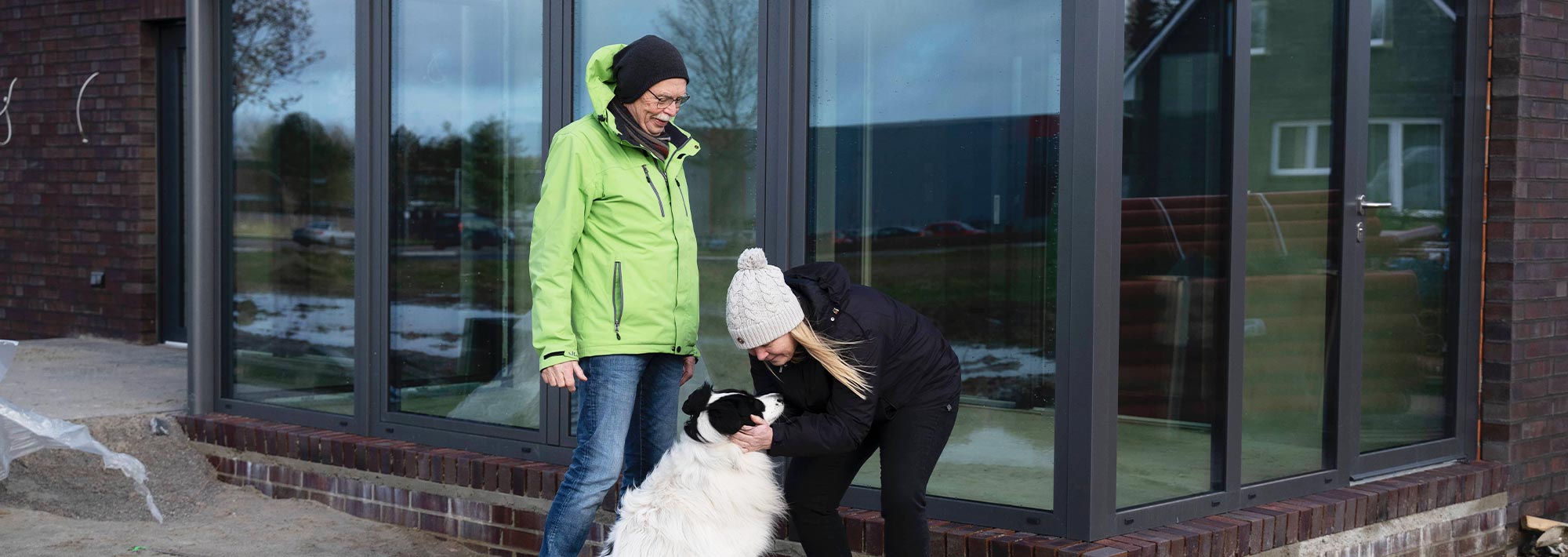 Ein Mann und eine Frau in Winterjacken und Mützen stehen vor einem modernen Glasgebäude, lächeln und streicheln einen großen schwarz-weißen Hund, als ob sie zurück zum Anfang gekommen wären.