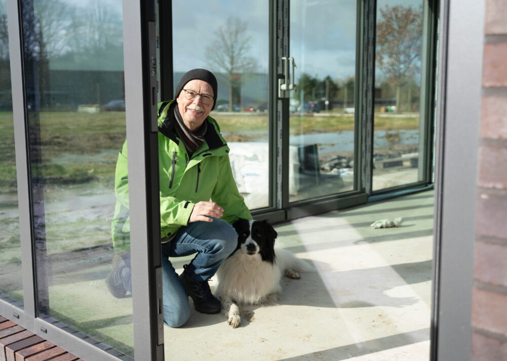 Eine ältere Person in grüner Jacke kniet lächelnd neben einem schwarz-weißen Hund in einem gläsernen Raum, der zeigt, dass Nähe mehr möglich macht. Durch die großen Fenster sind draußen Bäume und Autos zu sehen.