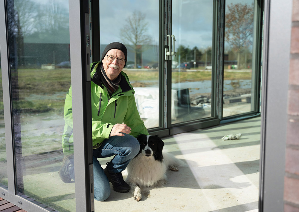 Eine Person mit grüner Jacke und schwarzem Hut kniet neben einem schwarz-weißen Hund in einem gläsernen Gebäude, in das das Sonnenlicht einfällt, als würde es sie zum Anfang zurückführen, während im Hintergrund eine Szene im Freien zu sehen ist.