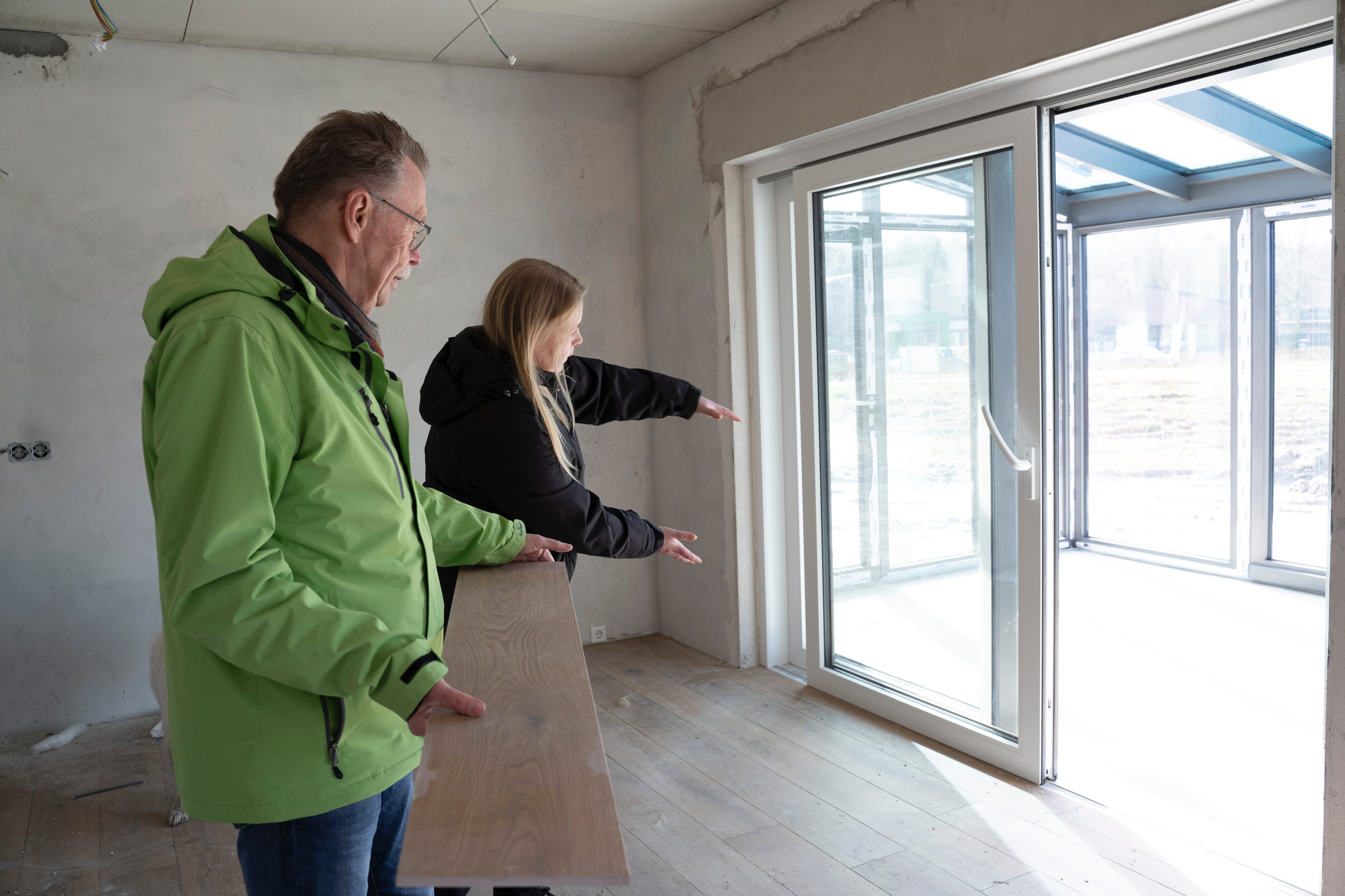 Zwei Menschen stehen in einem halbfertigen Raum; einer hält ein Holzbrett in der Hand, während der andere mit Gesten auf große Glastüren zugeht - Nähe macht mehr möglich -, während sie über Renovierungs- oder Baupläne diskutieren.
