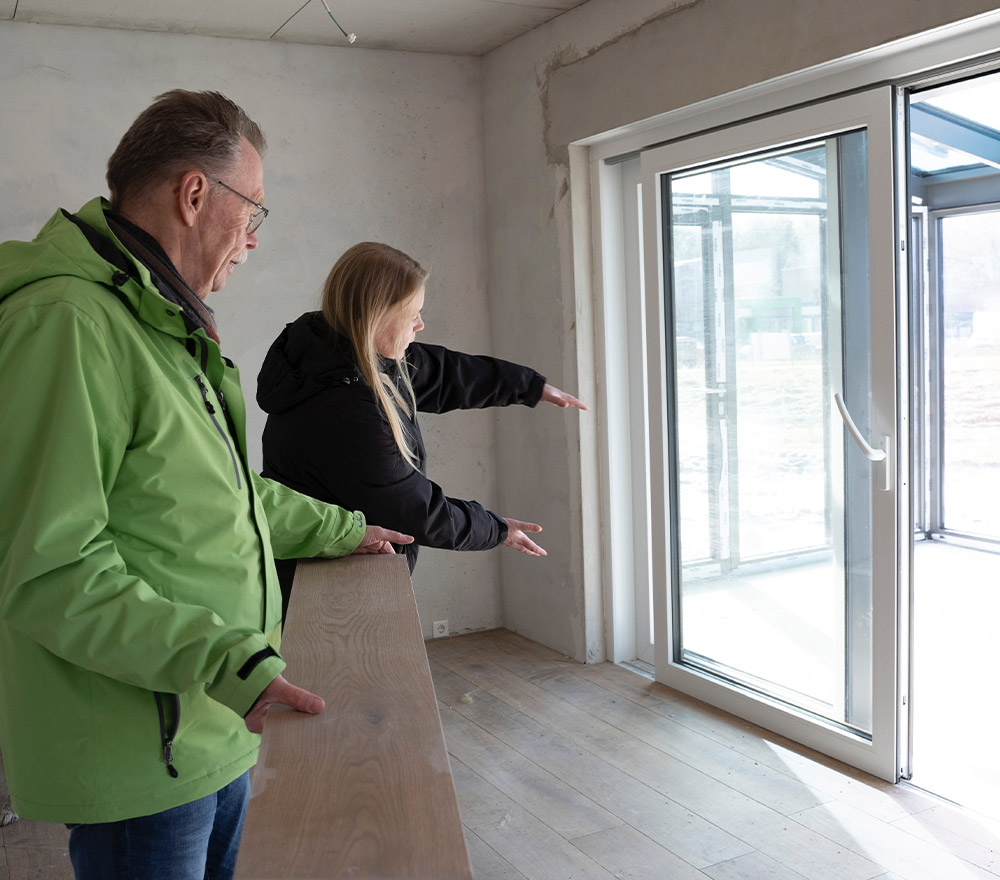 Zwei Personen in Jacken stehen in einem halbfertigen Raum mit nackten Wänden und Holzböden und zeigen auf große Glastüren, die sich nach draußen öffnen - bereit für einen Neuanfang, Zurück zum Anfang.