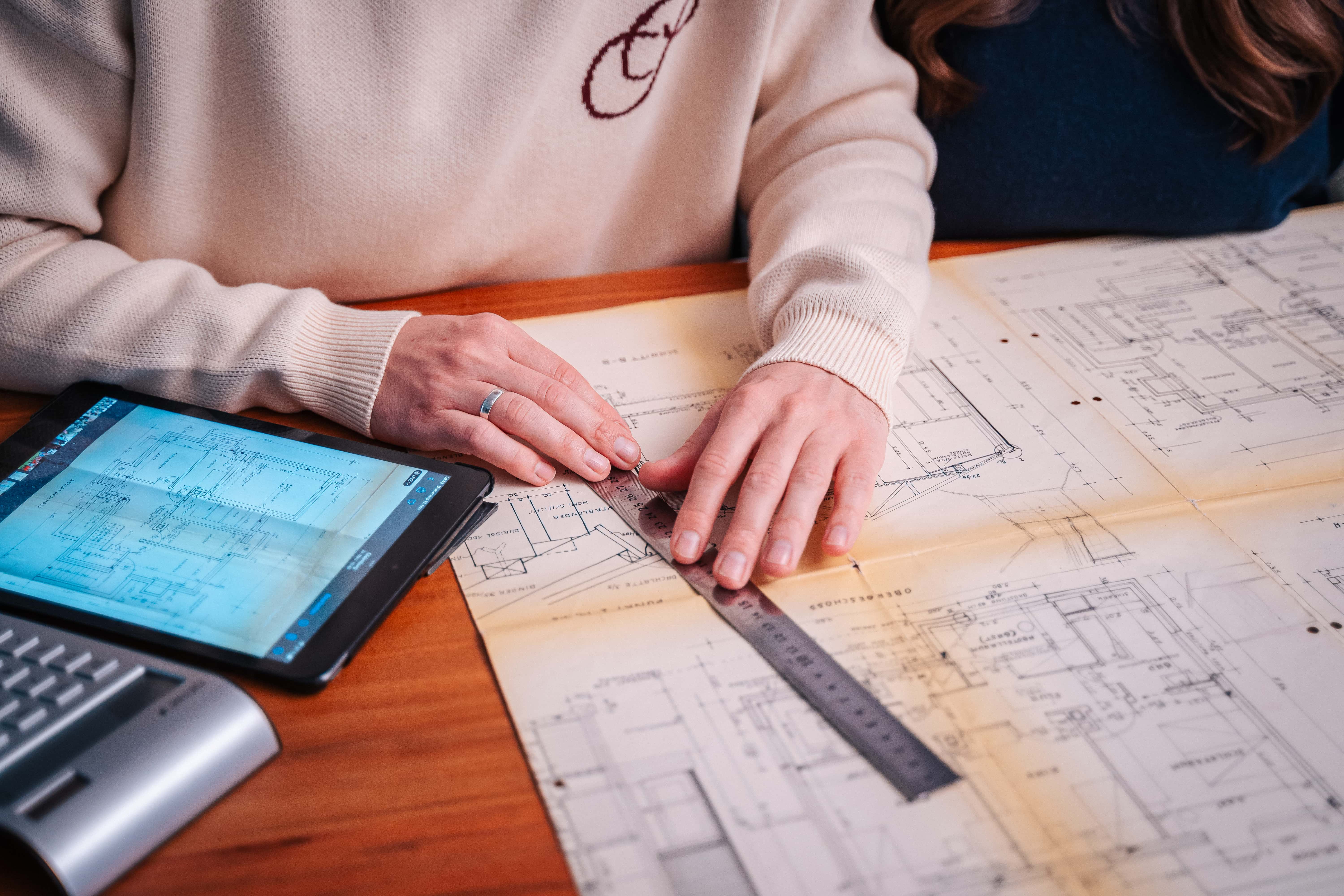 Zwei Personen prüfen auf einem Schreibtisch architektonische Entwürfe. Einer benutzt ein Lineal, um zu messen, während in der Nähe ein Tablet mit ähnlichen Plänen und eine Tastatur stehen. Nur ihre Hände und ein Teil ihrer Arme sind sichtbar und verkörpern den Ansatz "Zurück zum Anfang" des Designs.