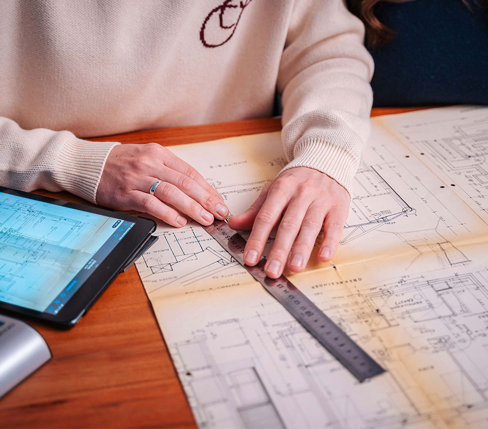 Eine Person misst mit einem Metalllineal und zeichnet Linien auf Architekturpläne, während ein Tablet mit ähnlichen Plänen in der Nähe auf dem hölzernen Schreibtisch steht - bereit, zurück zum Anfang zu gehen, falls Änderungen erforderlich sind.