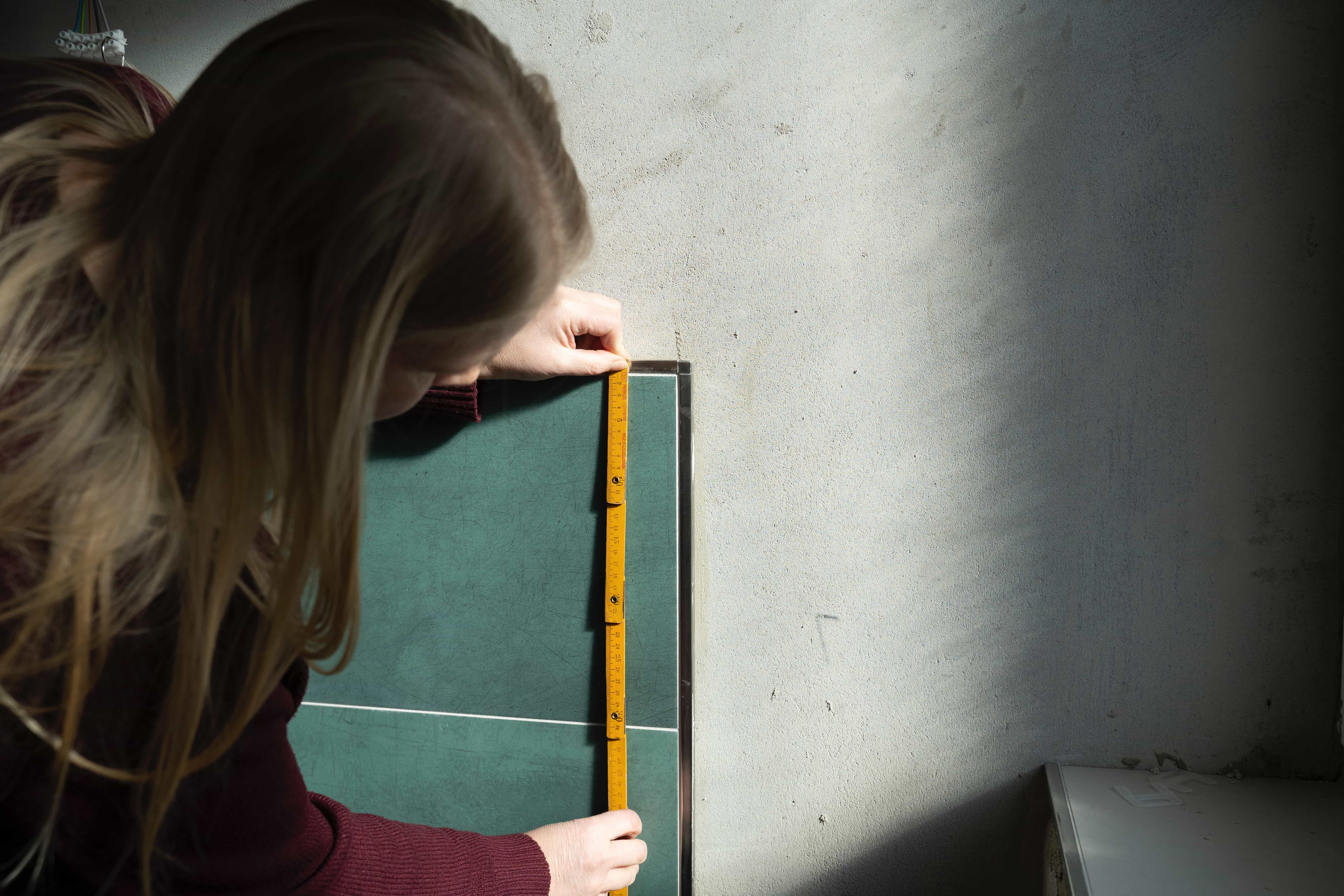 Eine Person mit langen Haaren misst mit einem gelben Maßband die Kante einer grünen Keramikfliese an einer hellgrauen Wand in natürlichem Sonnenlicht.