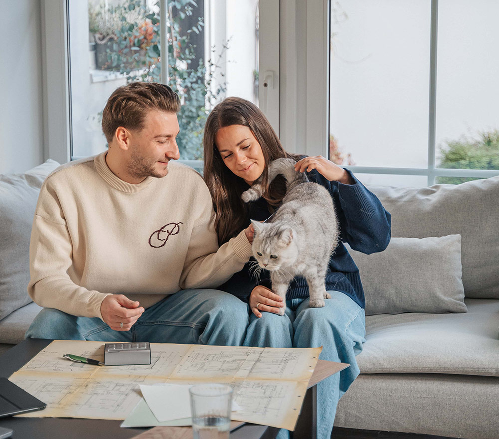 Ein Mann und eine Frau sitzen auf einer Couch, vor ihnen auf dem Tisch liegen Hauspläne. Die Frau streichelt eine graue Katze auf ihrem Schoß, während das Sonnenlicht durch das Fenster hereinströmt und eine gemütliche "Zurück zum Anfang"-Atmosphäre schafft, die an einen Neuanfang erinnert.