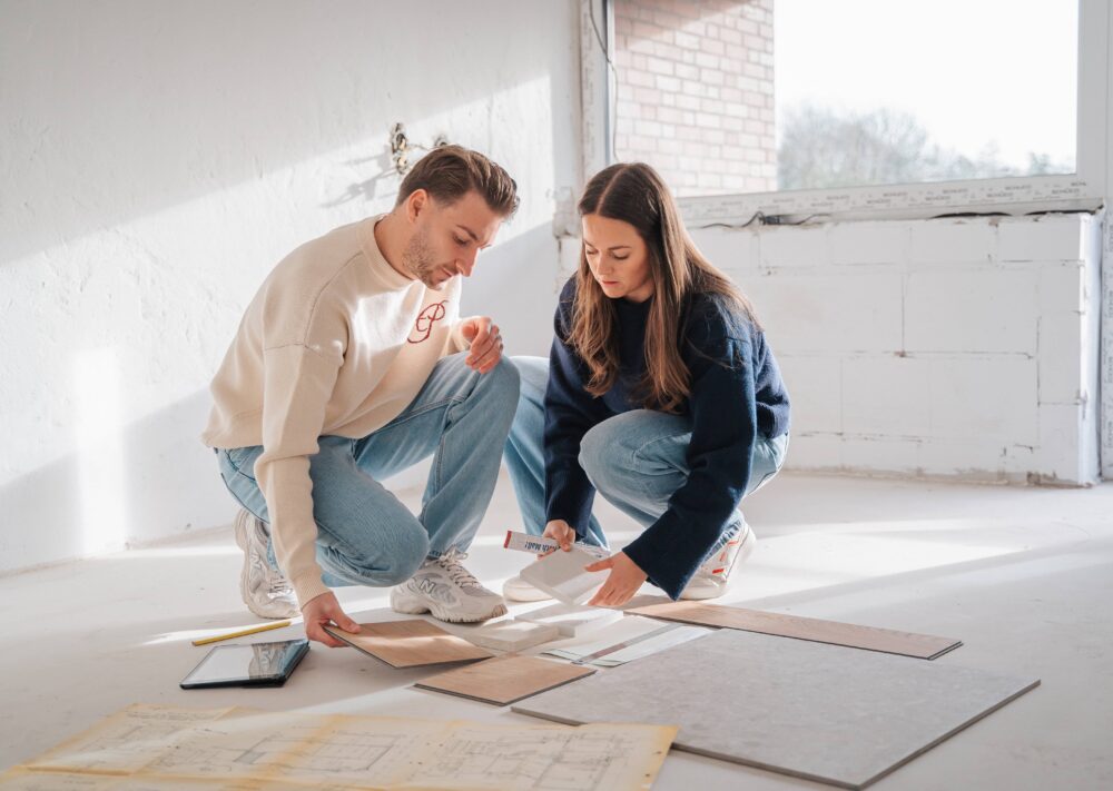 Ein Mann und eine Frau knien auf dem Boden eines hellen, unfertigen Raums und begutachten Bodenbelagsproben und Papiere. Umgeben von architektonischen Plänen und einem Tablet diskutieren sie Renovierungsoptionen - bereit für einen Neuanfang, zurück zum Anfang.