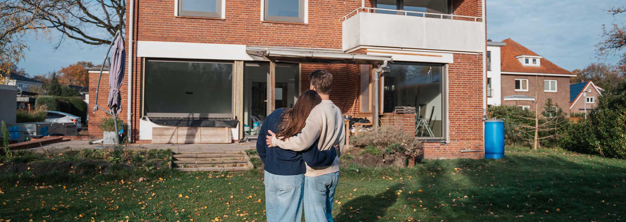 Ein Paar steht Arm in Arm auf dem Rasen vor einem zweistöckigen Backsteinhaus mit großen Fenstern und einer teilweise offenen Terrasse - bereit für ein neues Kapitel, das sich anfühlt wie Zurück zum Anfang.