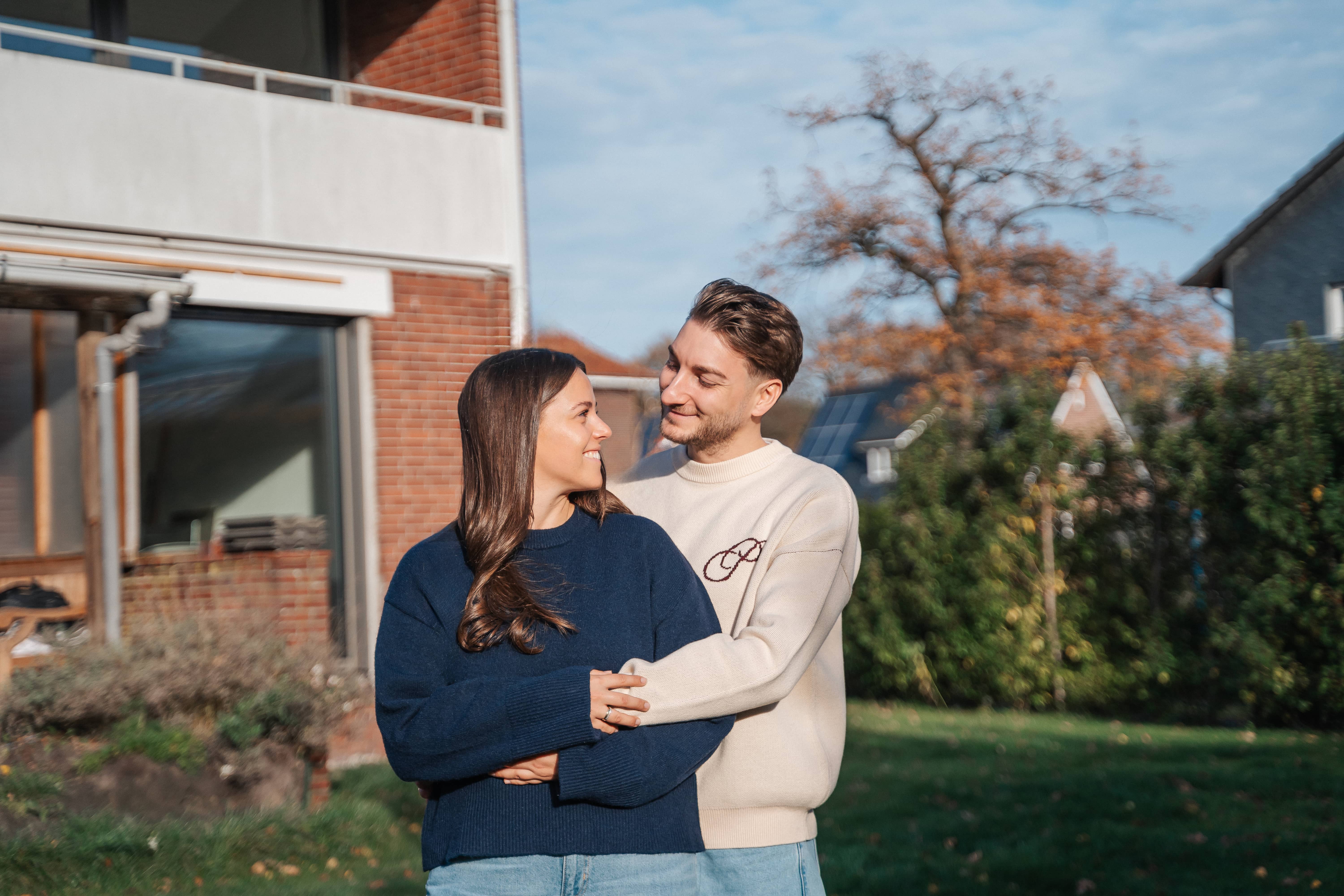 Ein Paar steht draußen in einem Garten, lächelt und umarmt sich, während es sich in die Augen schaut - Momente, die sich wie Zurück zum Anfang anfühlen. Sie sind leger gekleidet, mit einem Haus und Bäumen im Hintergrund an einem sonnigen Tag.