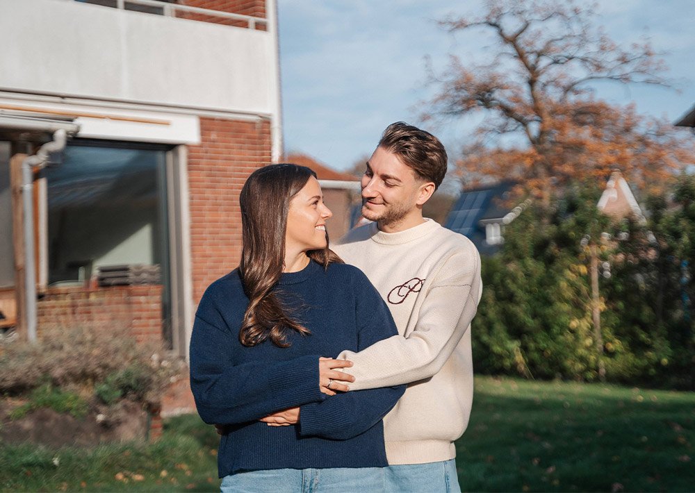 Ein junges Paar steht draußen in einem Garten, lächelt und umarmt sich gegenseitig. Sie tragen lässige Pullover und Jeans und strahlen vor Glück - ein perfekter Zurück-zum-Anfang-Moment mit einem Haus und Bäumen im Hintergrund.