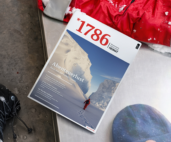 Eine Zeitschrift mit dem Titel 1786 liegt auf einem Tisch neben einem blauen Hut und einer roten Jacke. Das Titelbild des 1786 Online Beitrags zeigt eine Person, die durch tiefen Schnee zwischen hohen eisigen Klippen wandert, und trägt die deutsche Überschrift Abenteuerlust.
