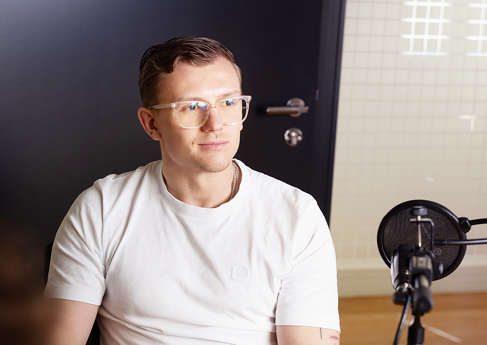 Ein Mann mit Klarsichtbrille und weißem T-Shirt sitzt vor einem schwarzen Mikrofon und blickt leicht zur Seite. Er befindet sich in einem modernen Innenraum mit einer schwarzen Tür und einer Glasscheibe, die an ein ZDF-Theater-Set erinnert.