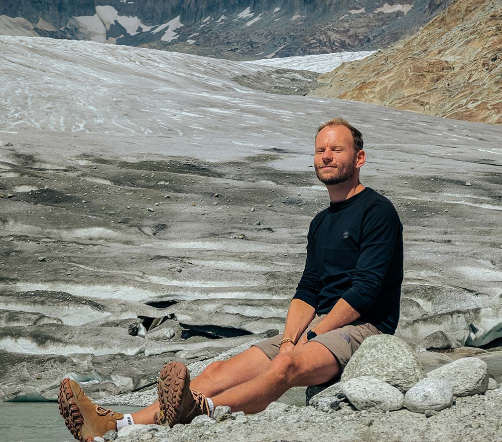 Ein Mann sitzt auf einem Felsen vor einem Gletscher, die Augen geschlossen und das Gesicht der Sonne zugewandt, und macht eine Pause auf seiner Fahrrad-Weltreise. Er trägt Wanderschuhe, kurze Hosen und ein langärmeliges Hemd; im Hintergrund erheben sich Schnee und felsige Berge.