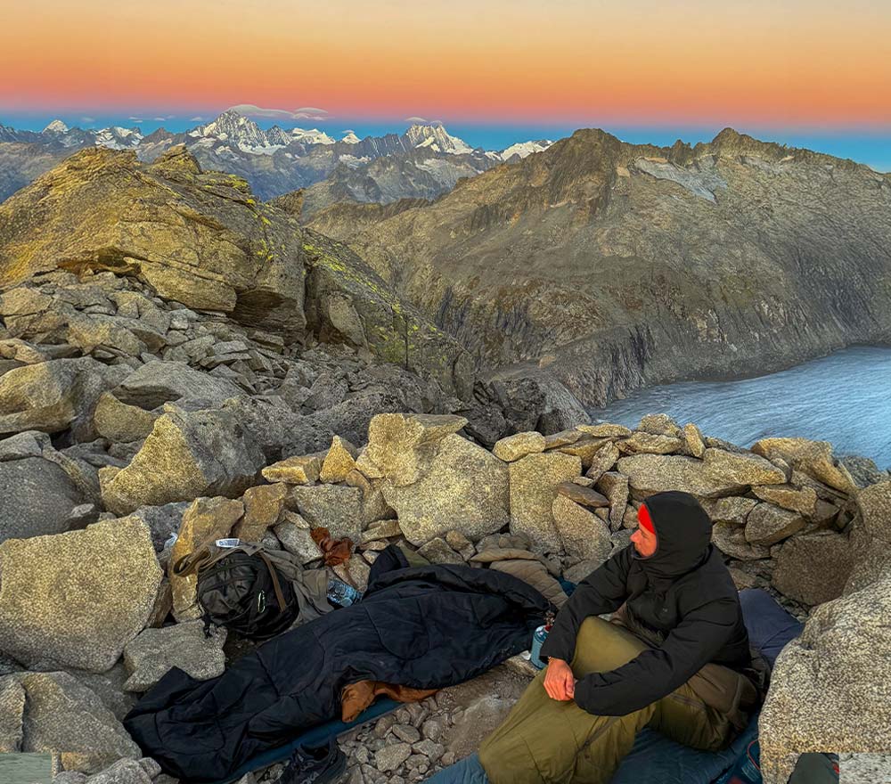 Eine Person in Outdoor-Ausrüstung sitzt bei Sonnenaufgang zwischen Felsen und Campingausrüstung auf einem Bergkamm und blickt auf ferne schneebedeckte Gipfel und einen See - eine atemberaubende Szene ihrer Fahrrad-Weltreise unter einem farbenfrohen Himmel.