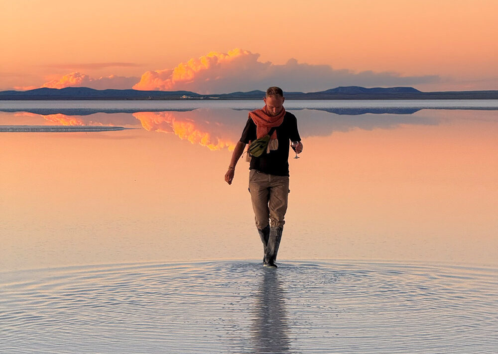 Eine Person auf einer Fahrrad Weltreise geht bei Sonnenuntergang durch seichtes Wasser und erzeugt Wellen. Der Himmel und die Wolken spiegeln sich auf der Wasseroberfläche, und in der Ferne sind die Hügel unter einem sanften orange-rosa Himmel zu sehen.