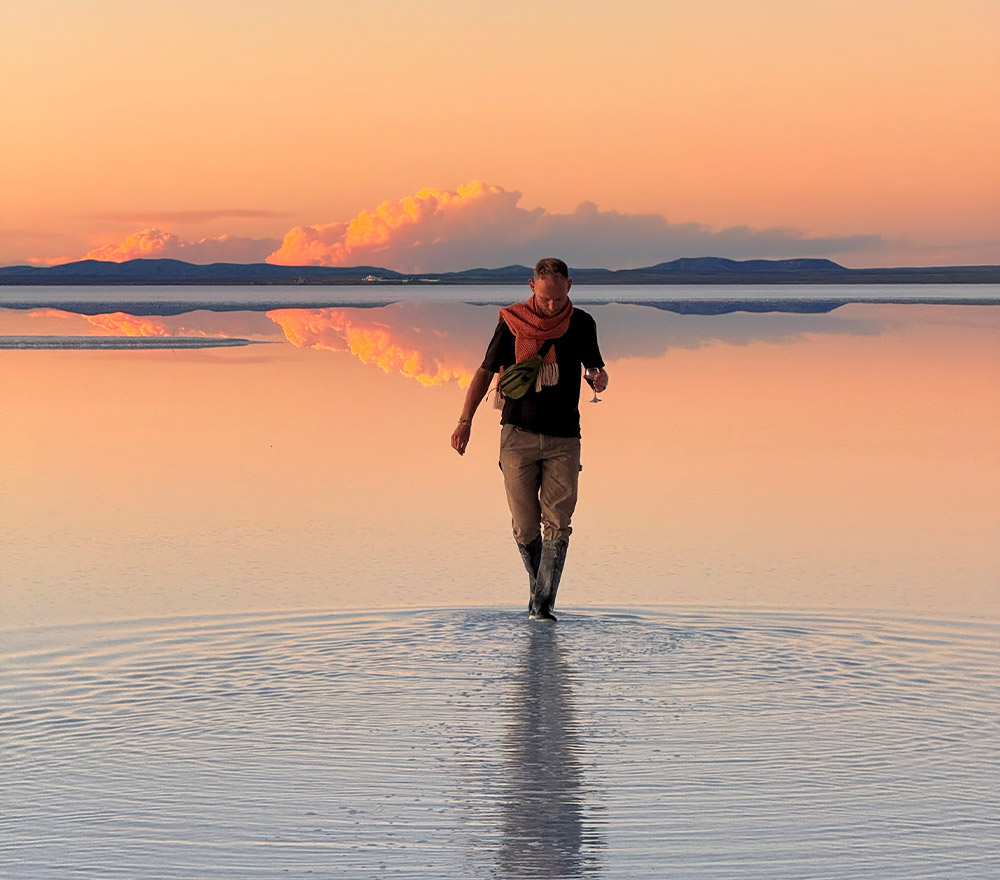 Eine Person auf einer Fahrrad Weltreise geht bei Sonnenuntergang durch seichtes Wasser und erzeugt Wellen. Der Himmel und die Wolken spiegeln sich auf der Wasseroberfläche, und in der Ferne sind die Hügel unter einem sanften orange-rosa Himmel zu sehen.