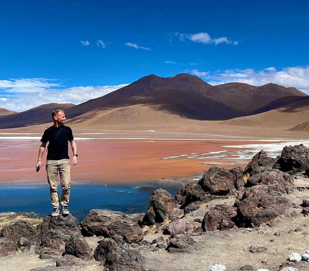 Ein Mann in schwarzem Hemd und beiger Hose steht während seiner Fahrrad Weltreise auf felsigem Gelände und blickt zur Seite. Im Hintergrund sind eine bunte Lagune, Berge und ein strahlend blauer Himmel zu sehen.