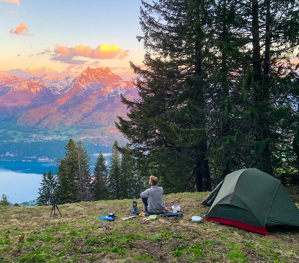 Eine Person auf einer Fahrrad Weltreise sitzt in der Nähe eines grünen Zeltes zwischen Bäumen auf einem grasbewachsenen Hügel und blickt auf eine malerische Berglandschaft mit einem See und schneebedeckten Gipfeln bei Sonnenuntergang. Die Campingausrüstung ist in der Nähe auf dem Boden ausgebreitet.