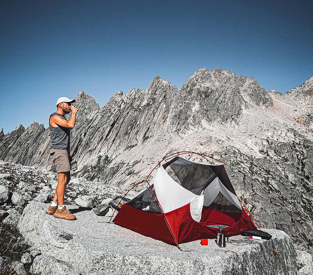 Ein Mann steht neben einem rot-grauen Zelt auf felsigem Gelände und trinkt aus einer Tasse. Campingausrüstung und ein Fahrrad Weltreise liegen in der Nähe verstreut, während sich im Hintergrund zerklüftete Berggipfel unter einem klaren blauen Himmel erheben.