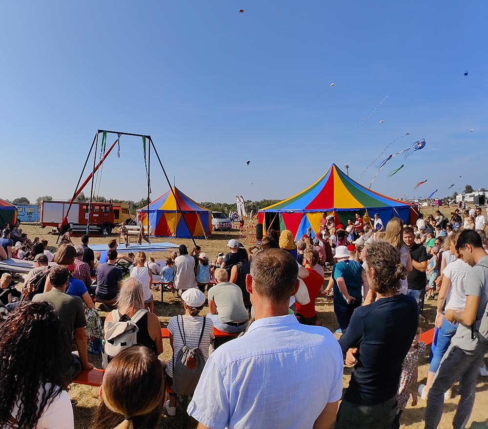Eine große Menschenmenge versammelt sich im Freien in der Nähe der bunten Zirkuszelte beim Drachenfest Lemwerder, um eine Vorstellung zu sehen. Drachen fliegen in den blauen Himmel, und die Menschen sitzen und stehen, um das Geschehen an einem sonnigen Tag zu beobachten.