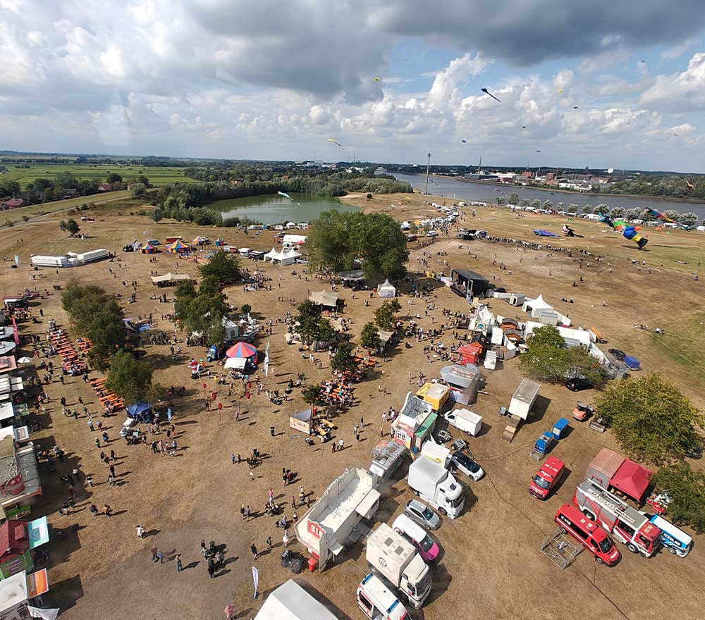 Die Luftaufnahme des Drachenfestes Lemwerder zeigt ein Freiluftfest mit Imbisswagen, Zelten, Menschen und Picknicktischen auf einer Wiese in der Nähe eines Teiches. Am teilweise bewölkten Himmel steigen Drachen auf, während an einer Seite Fahrzeuge geparkt sind.