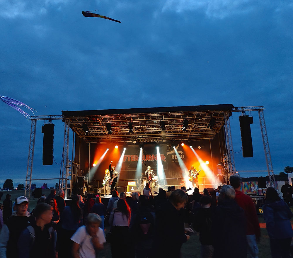 Eine Band spielt in der Abenddämmerung auf einer Freilichtbühne unter orangefarbenen Lichtern und unterhält das lebhafte Publikum des Drachenfestes Lemwerder, während über ihnen ein Drachen in den Himmel steigt und zur lebendigen Atmosphäre des Festes beiträgt.