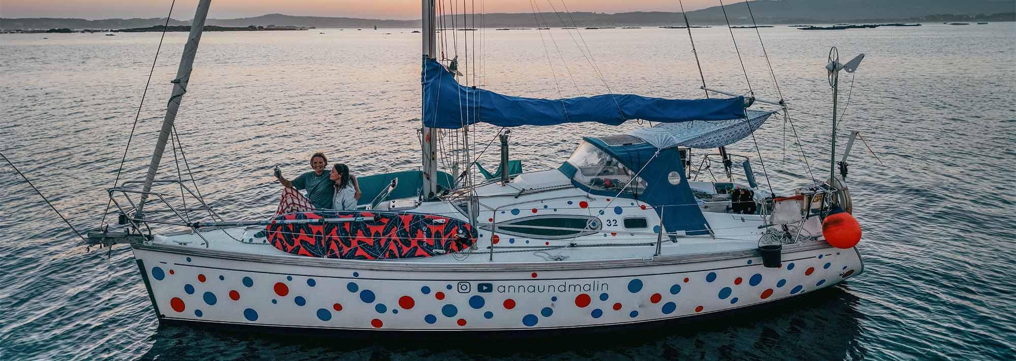 Ein weißes Segelboot mit roten und blauen Tupfen schwimmt bei Sonnenuntergang auf dem ruhigen Wasser. Anna und Malin stehen an Deck, lächeln und winken, bereit für ihr Segeln Abenteuer auf Weltreise. Der orangefarbene Himmel leuchtet, und am Horizont ist das ferne Land zu sehen.