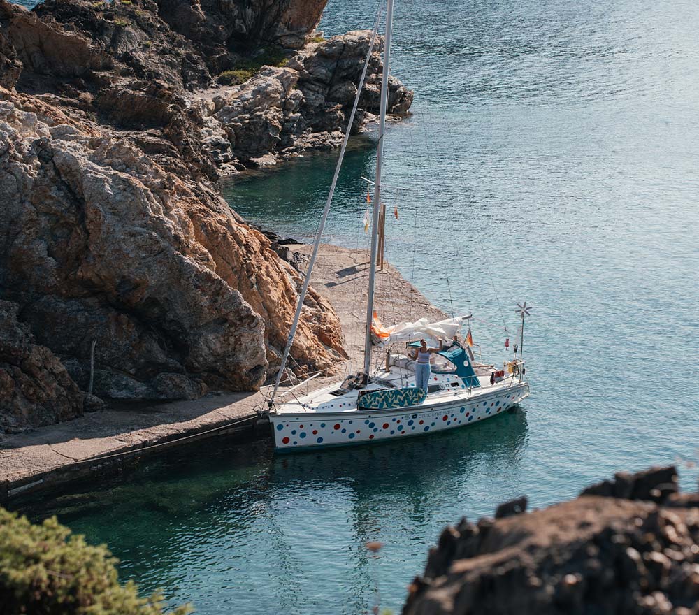 Ein weißes Segeln-Abenteuer-Segelboot mit bunten Tupfen liegt an einem felsigen Ufer auf klarem, blauem Wasser, im Hintergrund erheben sich im hellen Sonnenlicht schroffe Felsen - bereit für Anna Malins Weltreise.