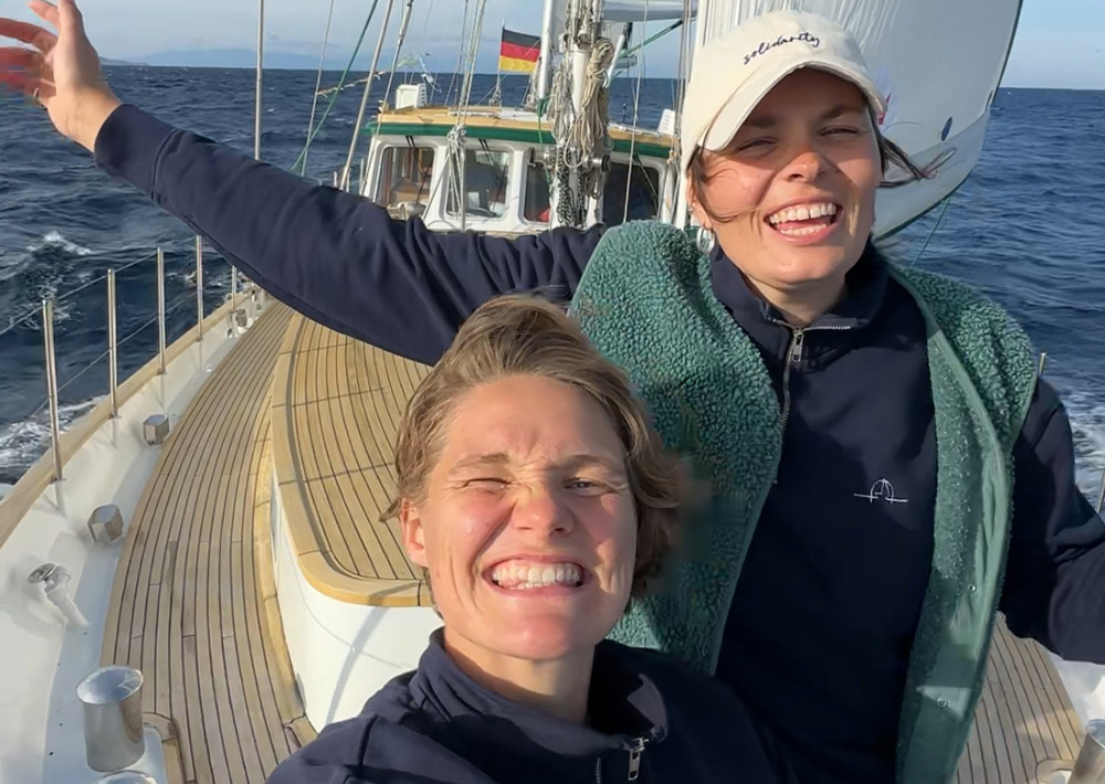 Zwei lächelnde Menschen auf dem Deck eines Segelbootes, die ihr Segeln Abenteuer genießen; einer steht mit weit ausgebreiteten Armen, während der andere in die Kamera zwinkert. Im Hintergrund: blauer Himmel, Meer und ein Boot mit deutscher Flagge - Anna Malins Weltreise in vollem Gange.