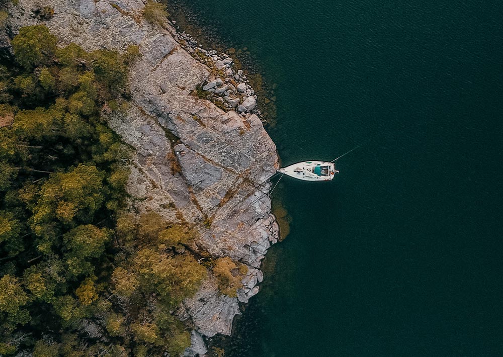 Ein weißes Segelboot liegt am felsigen Ufer vor Anker, umgeben von dichten Bäumen und tiefgrünem Wasser - ein heiterer Moment aus Anna Malins Segeln Abenteuer während ihrer Weltreise, wunderschön von oben eingefangen.
