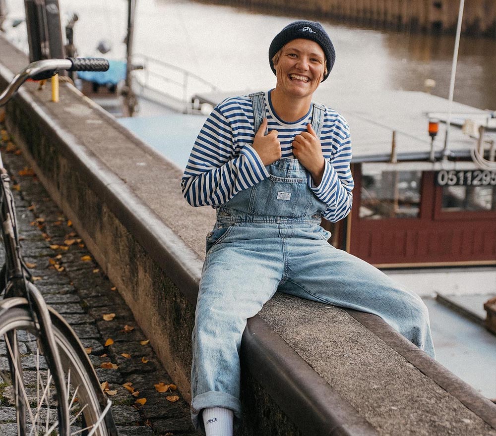 Eine lächelnde Person in einem gestreiften Hemd, einer Jeanslatzhose und einer Mütze sitzt auf einem Steinsockel am Flussufer und zeigt zwei Daumen nach oben - bereit für ein Segeln Abenteuer. Im Hintergrund sind ein Boot und ein Fahrrad zu sehen.