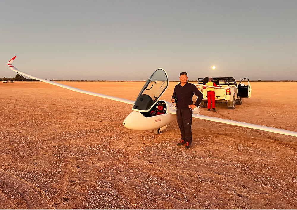 Eine Person steht neben einem Segelflugzeug und umarmt die Freiheit des Segelfliegens auf flachem, sandigem Gelände bei Sonnenuntergang. Eine andere Person befindet sich in der Nähe eines weißen Fahrzeugs im Hintergrund, unter einem klaren Himmel, an dem die Sonne oder der Mond tief steht.